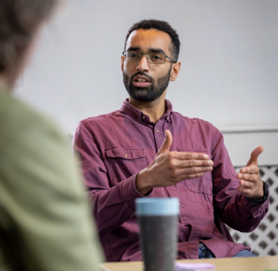 Image is of a man sitting down and having a discussion with a group. He is in the middle of talking and has his hands in front of him while he is expressing something. The person he is taking to has his back to the camera. There is a table between them and a reusable cup on the table..