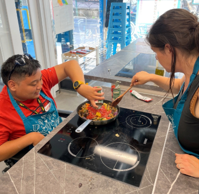 Image is of a boy in a wheelchair cooking in the the Brighton and Hove community kitchen - he is attending a cookery class. He is adding a tin of tomatoes into a pan on the the hob. The tutor is supporting him by stirring the ingredients in the pan. 